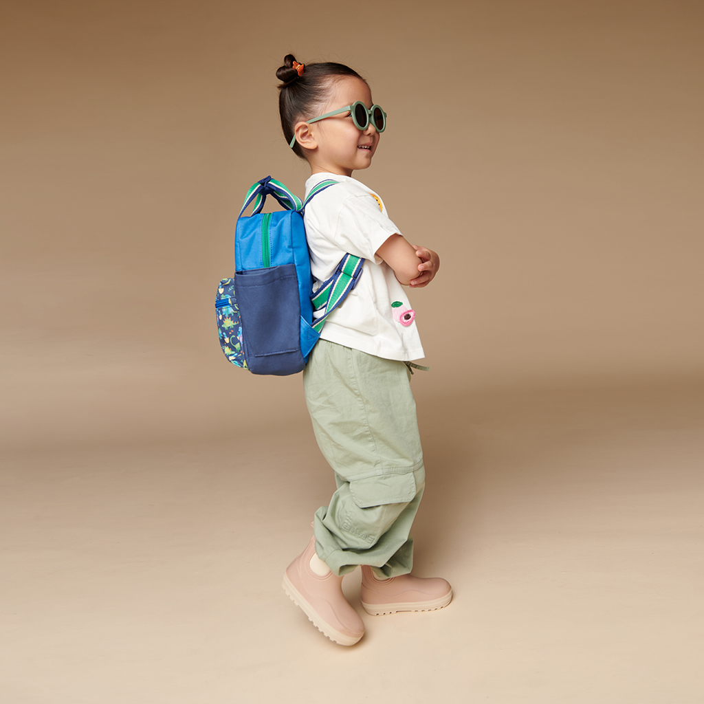 101601542 Itzy Bitzy Bag™ Toddler Backpack - Raining Dinos (11) Young child with a top knot hairstyle, sunglasses, and a backpack, wearing a white shirt, green pants, beige boots, and the Itzy Ritzy Sweetie Strap Plus Bear Silicone Pacifier Clip, standing against a plain background.
