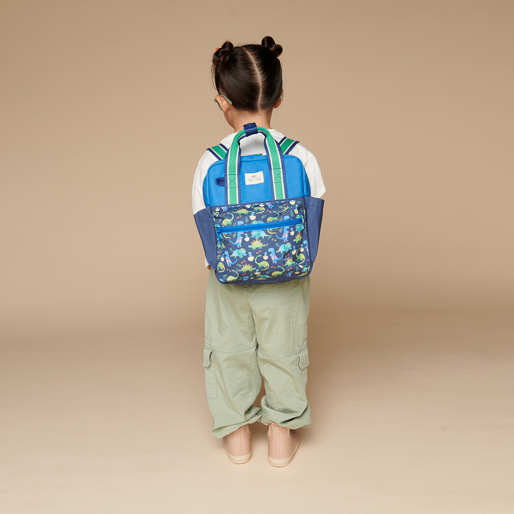 101601542 Itzy Bitzy Bag™ Toddler Backpack - Raining Dinos (12) A young child facing away, standing against a beige background, is wearing a blue backpack with a dinosaur pattern, green pants, a white shirt, pink shoes, and sporting an Itzy Ritzy Sweetie Strap Plus Bear Silicone Pacifier Clip.