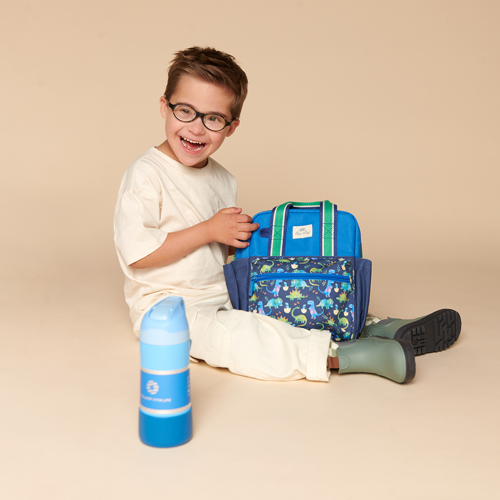 101601542 Itzy Bitzy Bag™ Toddler Backpack - Raining Dinos (13) A child with short hair and glasses sits on the floor, smiling and holding an Itzy Ritzy Sweetie Strap Plus Bear Silicone Pacifier Clip. A blue water bottle is placed in the foreground.