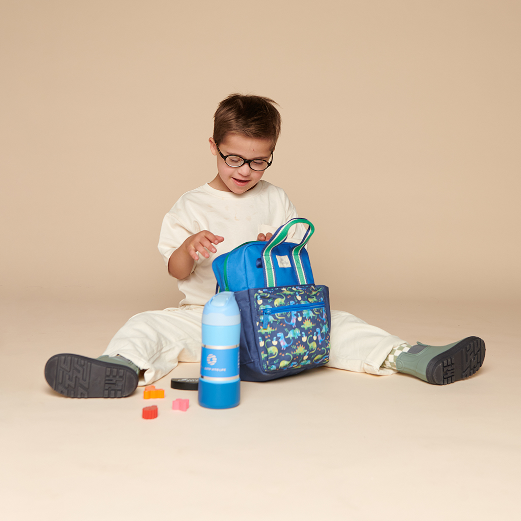 101601542 Itzy Bitzy Bag™ Toddler Backpack - Raining Dinos (14) A young child wearing glasses sits on the floor with legs spread, looking into a blue backpack. A water bottle and some small colorful blocks are placed in front, along with an Itzy Ritzy Sweetie Strap Plus Bear Silicone Pacifier Clip.