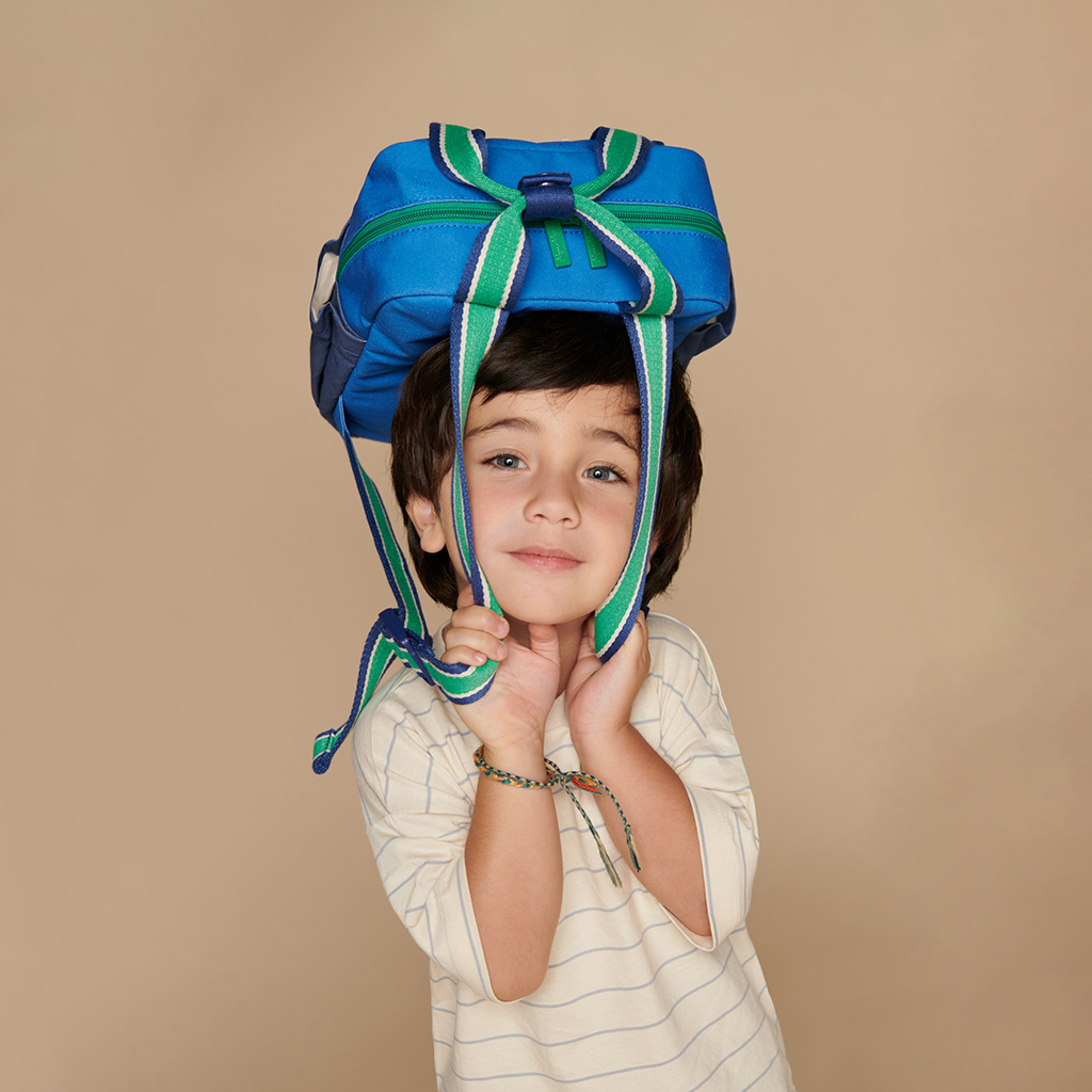 101601542 Itzy Bitzy Bag™ Toddler Backpack - Raining Dinos (17) A young boy with dark hair is wearing the Itzy Ritzy Sweetie Strap Plus Bear Silicone Pacifier Clip as a blue backpack on his head, with his hands holding the straps under his chin. He is dressed in a white and light blue striped shirt.