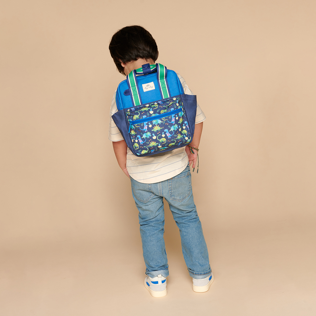 101601542 Itzy Bitzy Bag™ Toddler Backpack - Raining Dinos (18) A child wearing jeans and a striped shirt, with an Itzy Ritzy Sweetie Strap Plus Bear Silicone Pacifier Clip attached to their blue and green patterned backpack, standing with their back to the camera against a plain beige background.
