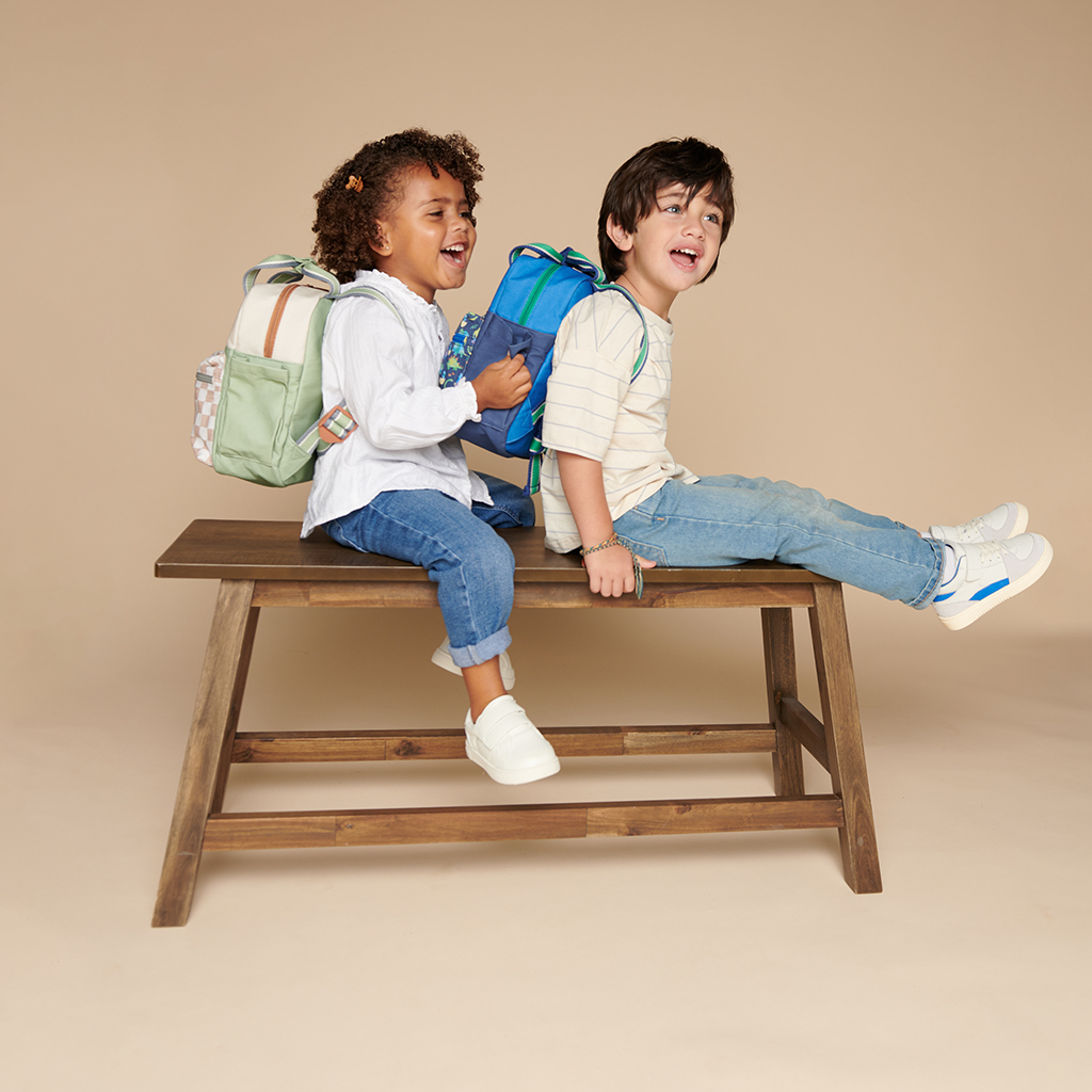 101601542 Itzy Bitzy Bag™ Toddler Backpack - Raining Dinos (20) Two children with backpacks sit and laugh on a wooden bench against a plain background. One child, wearing a white top, is using an Itzy Ritzy Sweetie Strap Plus Bear Silicone Pacifier Clip, while the other child wears a striped shirt.