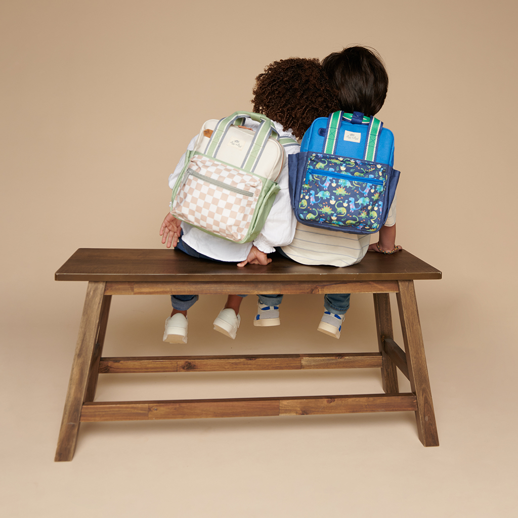 101601542 Itzy Bitzy Bag™ Toddler Backpack - Raining Dinos (3) Two children wearing colorful backpacks sit side by side on a wooden bench, both looking away from the camera, with one child holding an Itzy Ritzy Sweetie Strap Plus Bear Silicone Pacifier Clip.
