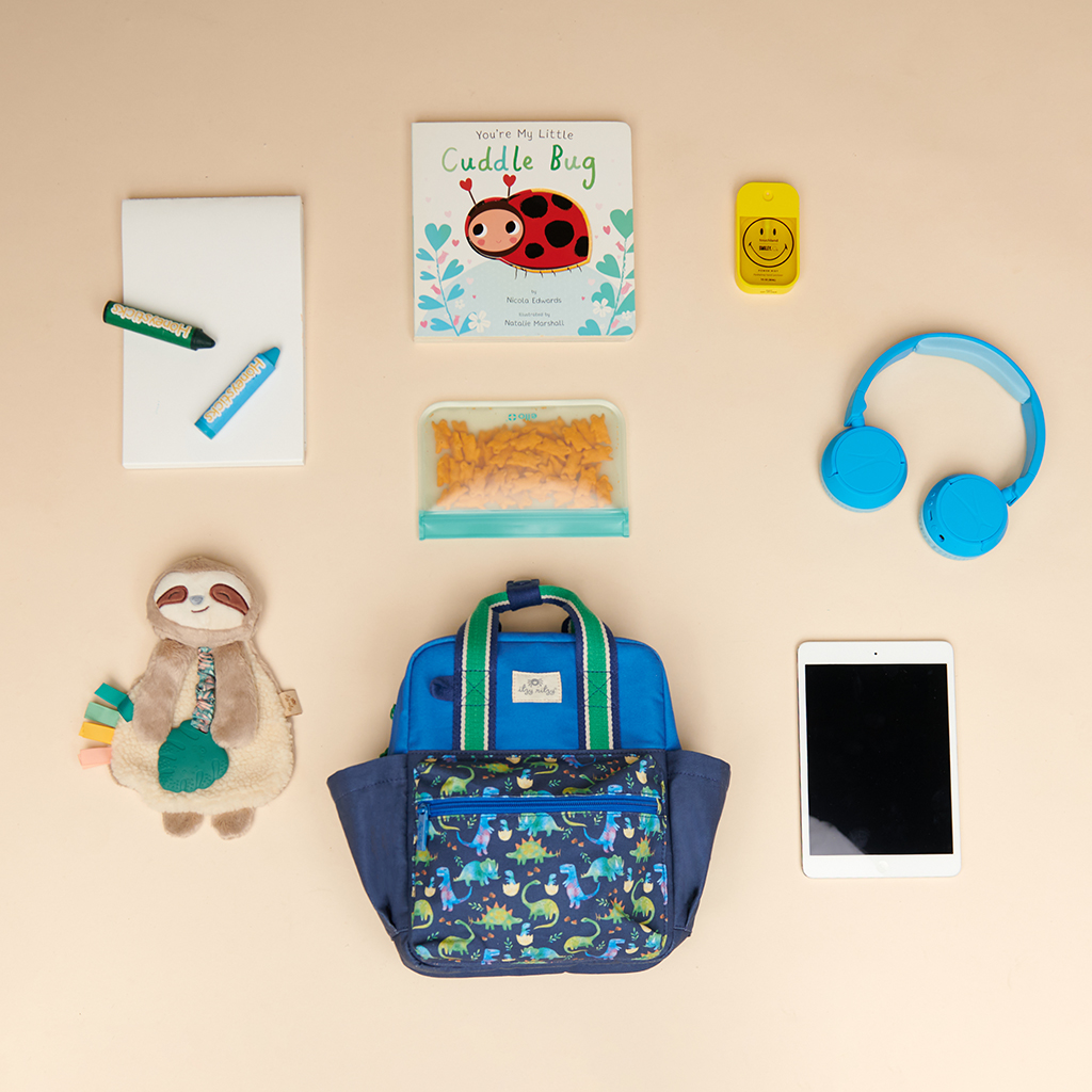 101601542 Itzy Bitzy Bag™ Toddler Backpack - Raining Dinos (8) A neatly arranged assortment featuring an Itzy Ritzy Sweetie Strap Plus Bear Silicone Pacifier Clip, a stuffed sloth, a book titled "You're My Little Cuddle Bug," headphones, tablet, notebook with pencil, lunchbox with snacks, smiley face toy, and child's backpack.