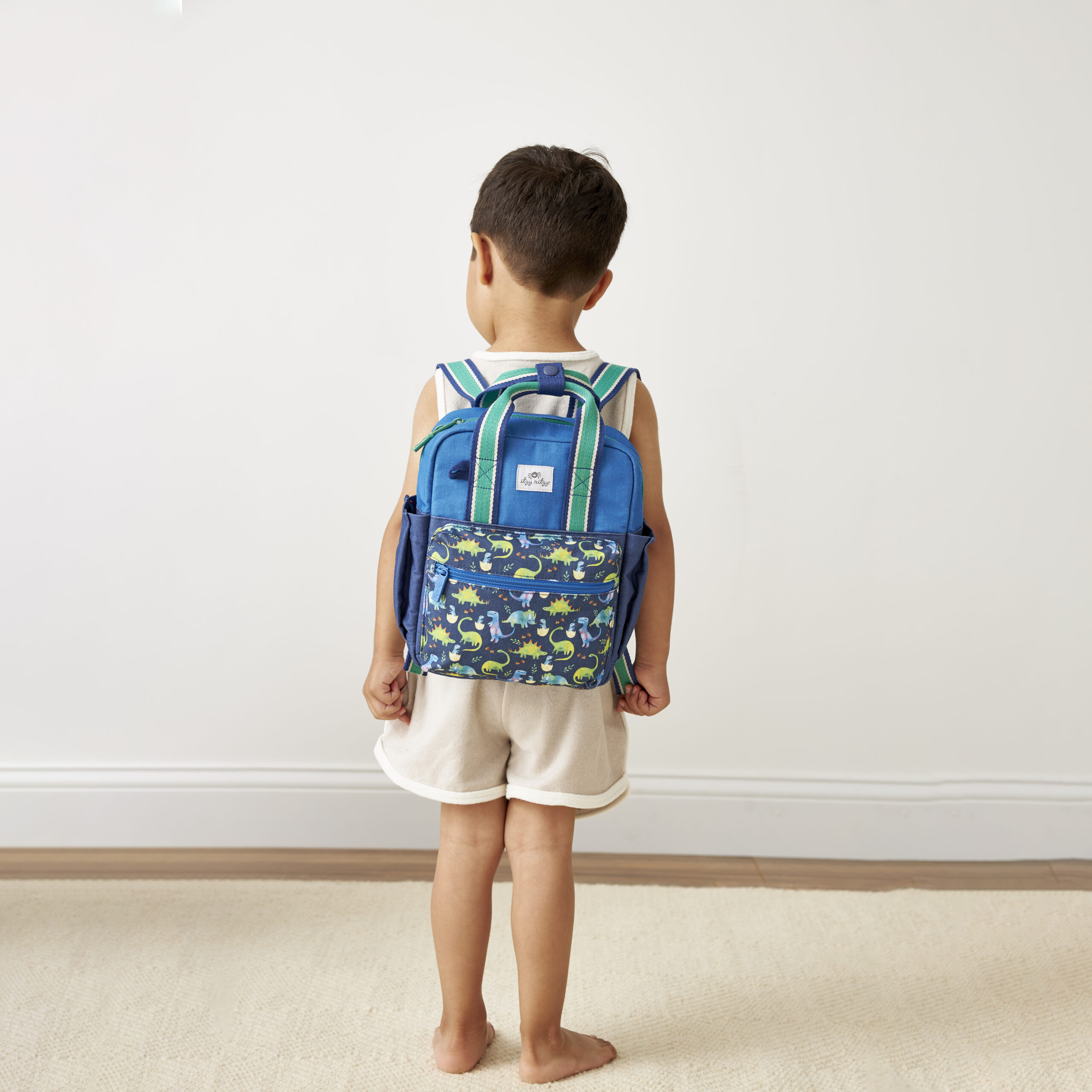 101601542 Itzy Bitzy Bag™ Toddler Backpack - Raining Dinos A young child with short hair stands facing away, wearing a white tank top, beige shorts, and a blue backpack with a floral pattern. The background features a plain wall and a light carpet. The child is accessorized with an Itzy Ritzy Sweetie Strap Plus Bear Silicone Pacifier Clip.