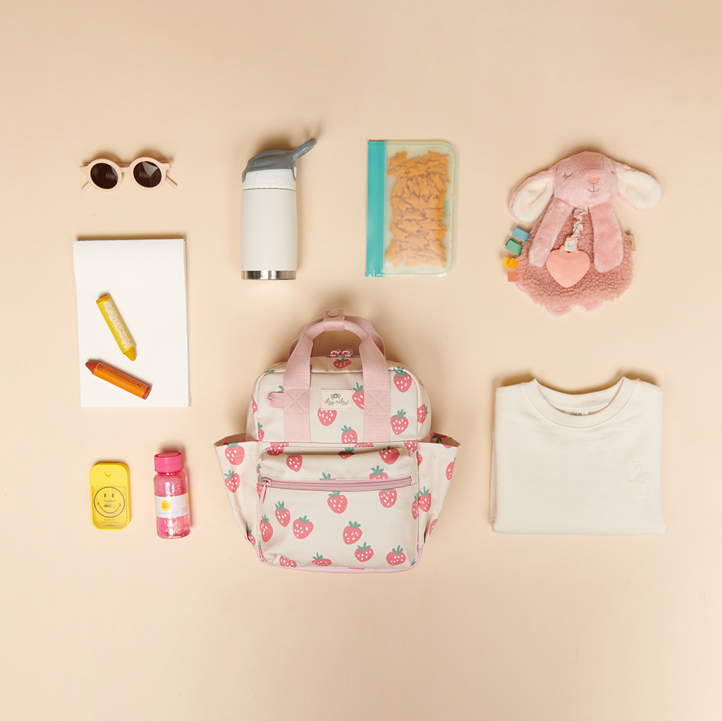 101601543 Itzy Bitzy Bag™ Toddler Backpack - Strawberries & Cream (1) A small backpack with a strawberry design is surrounded by sunglasses, a thermos, snacks in a bag, a toy bunny, a notebook, a crayon, lip balm, a snack container, and an Itzy Ritzy Sweetie Strap Plus Bear Silicone Pacifier Clip.
