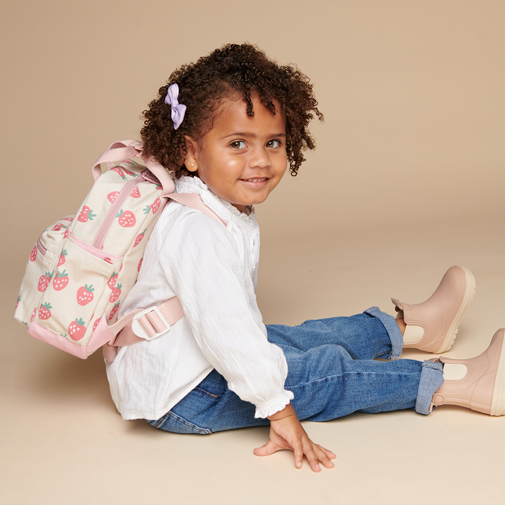 101601543 Itzy Bitzy Bag™ Toddler Backpack - Strawberries & Cream (10) A young child with curly hair, wearing a white shirt, jeans, and beige boots, sits on the floor with a strawberry-patterned backpack and a purple bow in their hair, accessorized with an Itzy Ritzy Sweetie Strap Plus Bear Silicone Pacifier Clip.