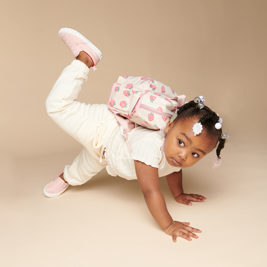 101601543 Itzy Bitzy Bag™ Toddler Backpack - Strawberries & Cream (11) A young child with a pink backpack performs a hands-and-knees pose against a beige background. She is wearing ivory-colored clothing, pink shoes, and an Itzy Ritzy Sweetie Strap Plus Bear Silicone Pacifier Clip.