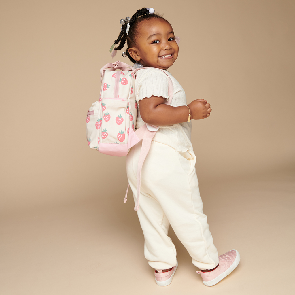 101601543 Itzy Bitzy Bag™ Toddler Backpack - Strawberries & Cream (12) A young child with braided hair smiles and poses sideways, wearing a white outfit, pink shoes, and carrying an adorable backpack with a strawberry pattern, accessorized perfectly with the Itzy Ritzy Sweetie Strap Plus Bear Silicone Pacifier Clip.