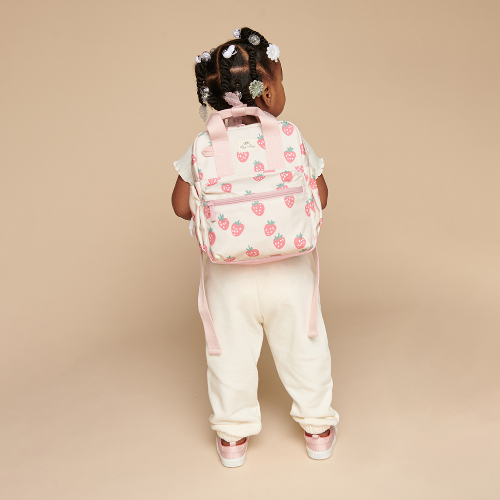 101601543 Itzy Bitzy Bag™ Toddler Backpack - Strawberries & Cream (13) A toddler wearing cream-colored attire stands facing away on a neutral background, showcasing a pink strawberry-patterned backpack and adorning their hairstyle with white and pink hair clips, along with an Itzy Ritzy Sweetie Strap Plus Bear Silicone Pacifier Clip.