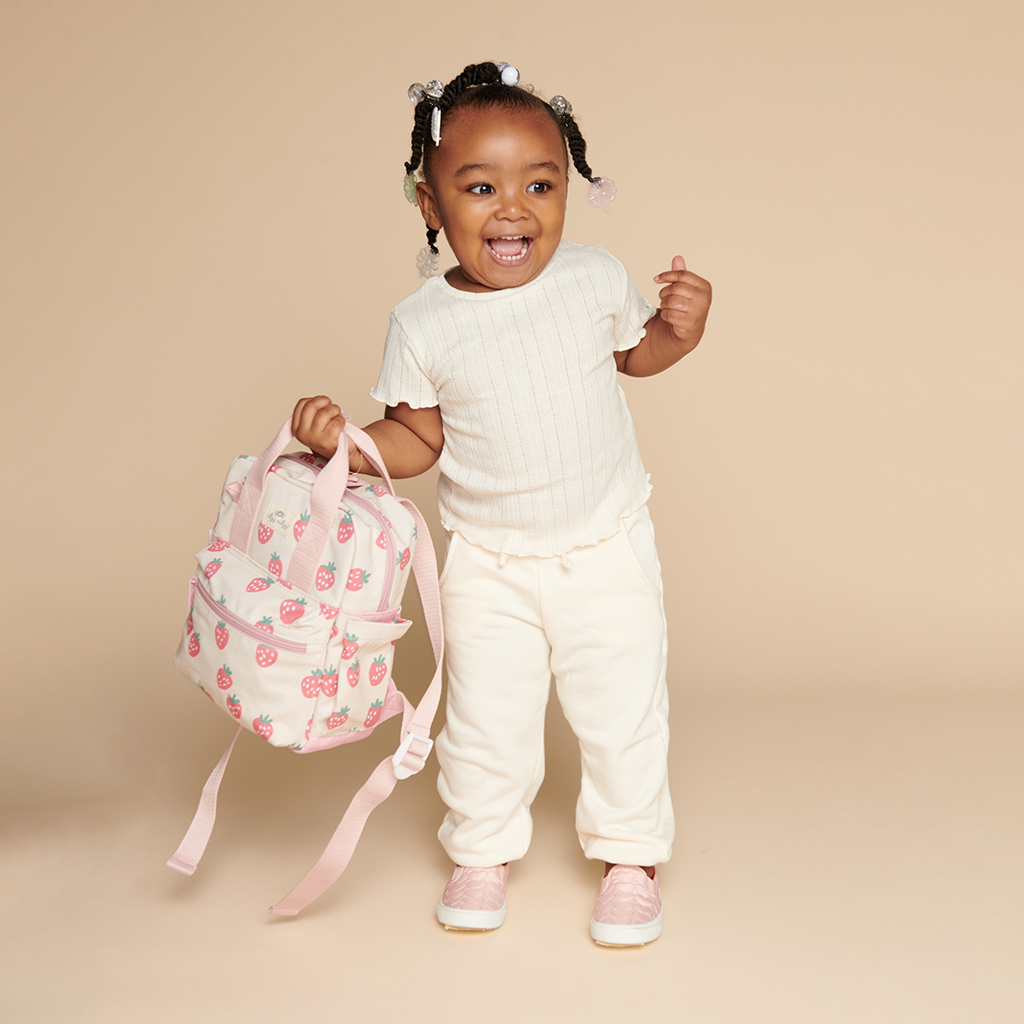 101601543 Itzy Bitzy Bag™ Toddler Backpack - Strawberries & Cream (14) A smiling toddler stands against a beige background, holding an Itzy Ritzy Sweetie Strap Plus Bear Silicone Pacifier Clip in one hand. The child wears a white shirt and pants, with pink shoes.