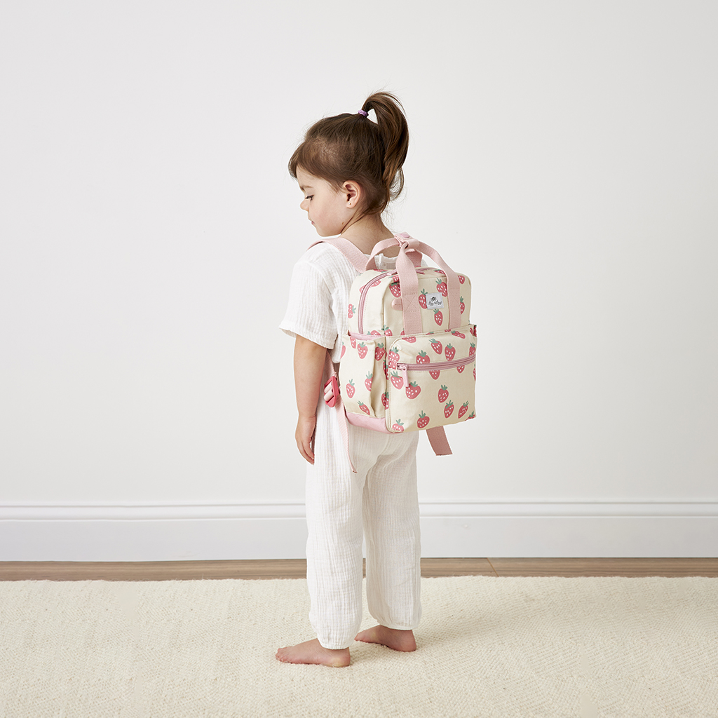 101601543 Itzy Bitzy Bag™ Toddler Backpack - Strawberries & Cream (16) A young child with light brown hair stands in profile, clad in an all-white outfit and carrying a pink strawberry-patterned backpack. She is barefoot on a light-colored rug against a white wall, with the Itzy Ritzy Sweetie Strap Plus Bear Silicone Pacifier Clip clipped to her attire.