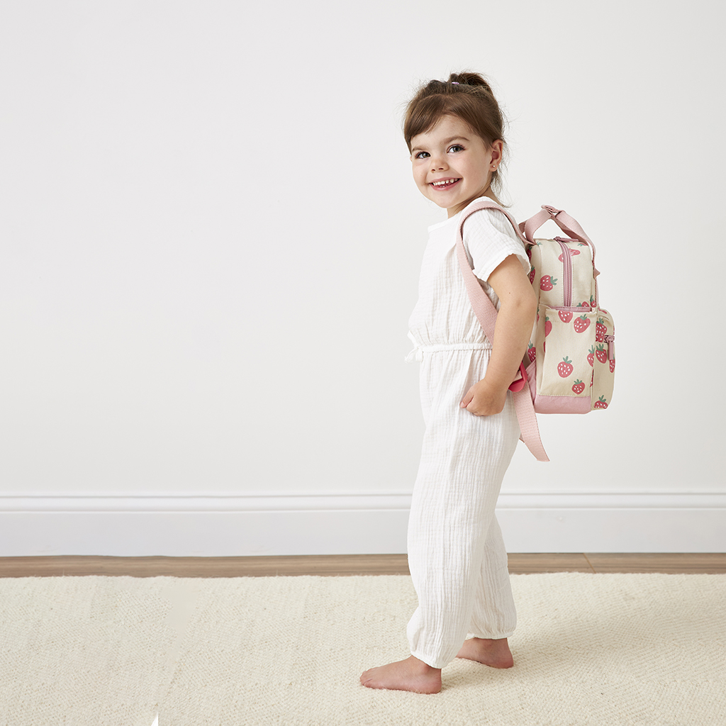 101601543 Itzy Bitzy Bag™ Toddler Backpack - Strawberries & Cream (17) A smiling young girl in white clothes stands sideways, holding an Itzy Ritzy Sweetie Strap Plus Bear Silicone Pacifier Clip over her shoulder against a white background.