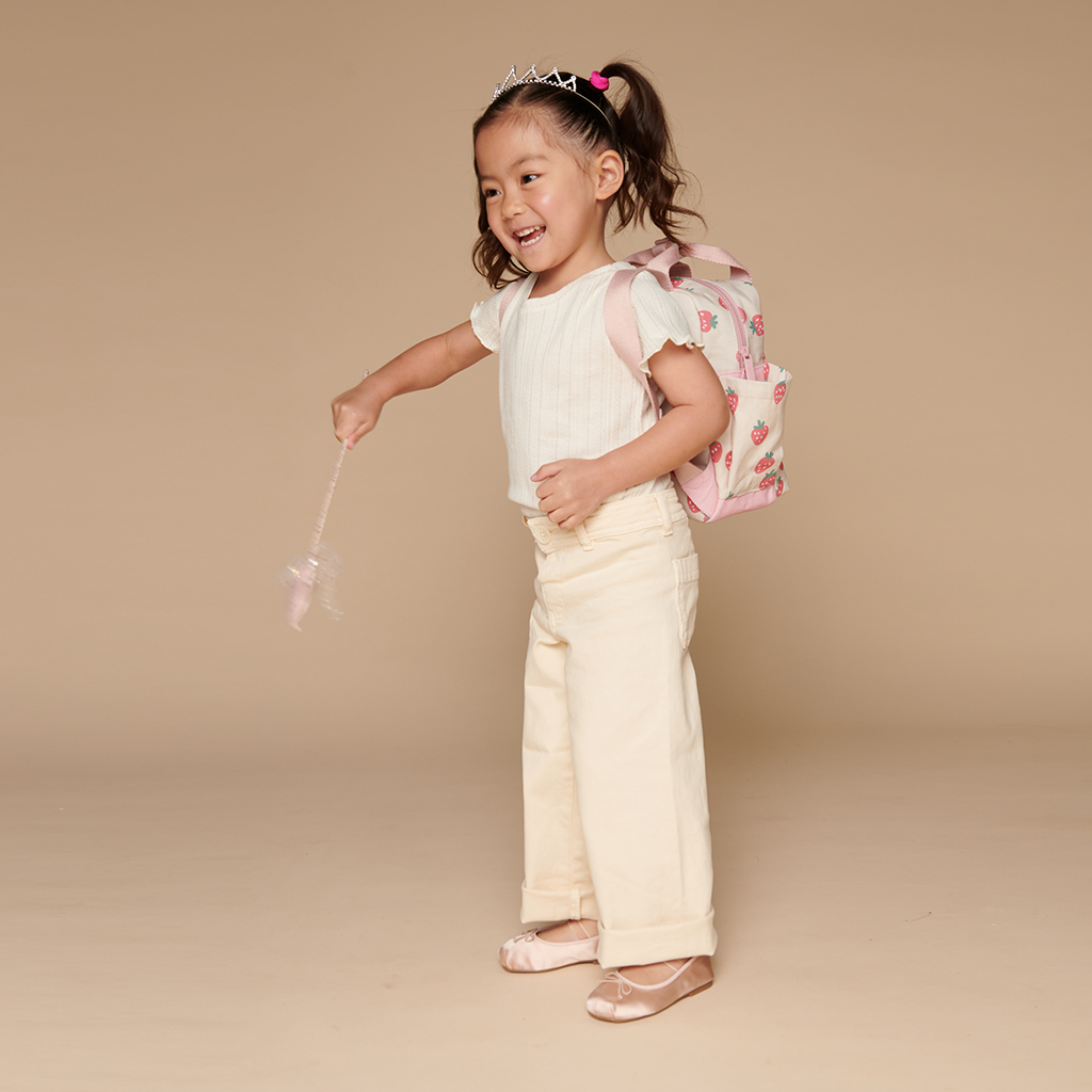 101601543 Itzy Bitzy Bag™ Toddler Backpack - Strawberries & Cream (8) A young girl smiling while wearing a tiara, white shirt, cream pants, and ballet shoes holds a wand with an Itzy Ritzy Sweetie Strap Plus Bear Silicone Pacifier Clip visible on her floral pink backpack against a beige background.