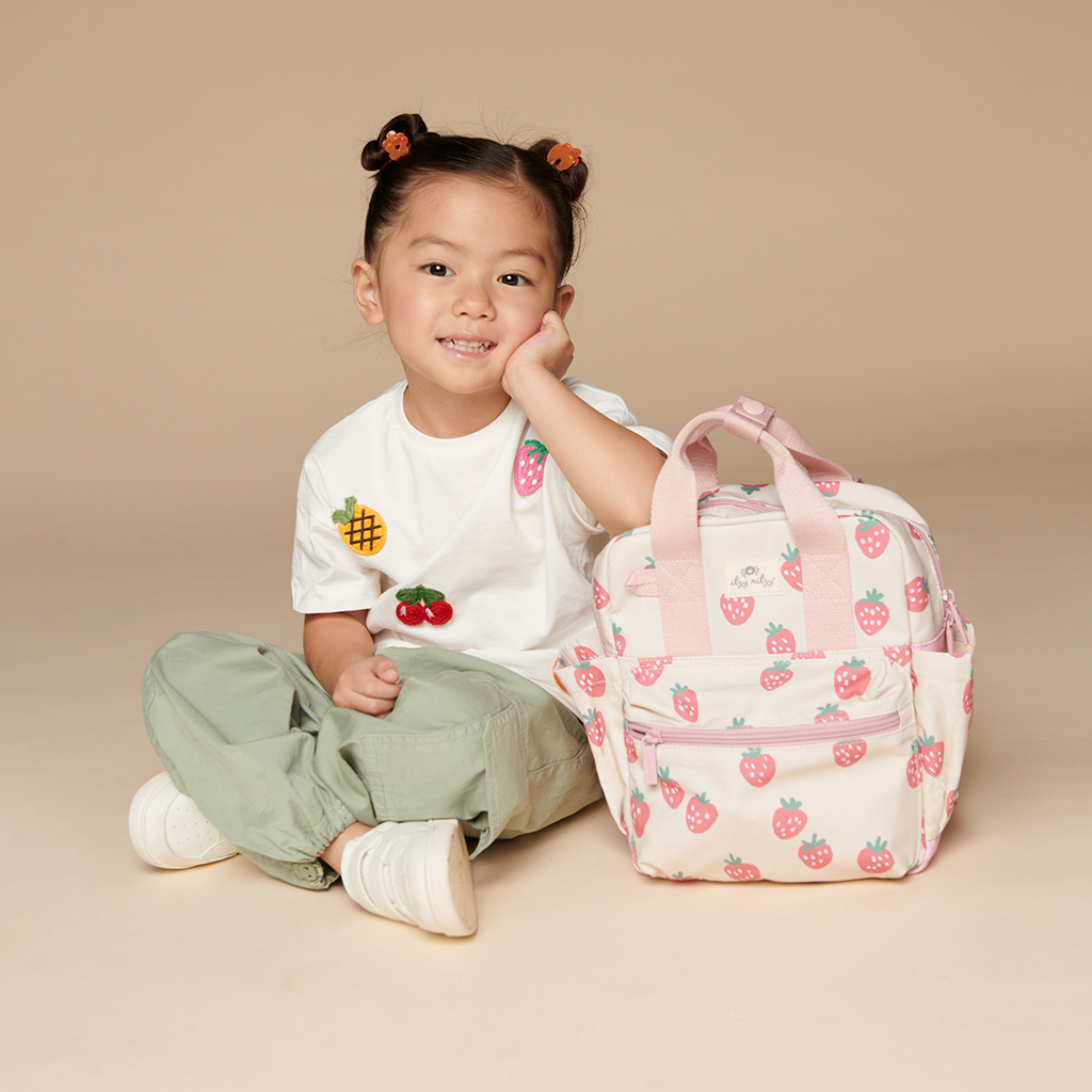 101601543 Itzy Bitzy Bag™ Toddler Backpack - Strawberries & Cream (9) A young child sits on the floor with a hand on their cheek, wearing a white shirt with fruit graphics, green pants, and white shoes, next to an Itzy Ritzy Sweetie Strap Plus Bear Silicone Pacifier Clip in pink featuring strawberry prints.