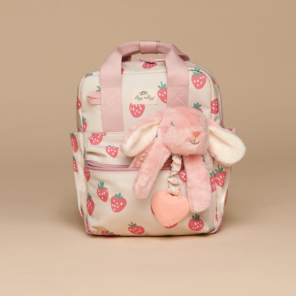 101601543 Itzy Bitzy Bag™ Toddler Backpack - Strawberries & Cream A small backpack with pink handles and strawberry patterns, featuring the Itzy Ritzy Sweetie Strap Plus Bear Silicone Pacifier Clip attached to the front pocket.