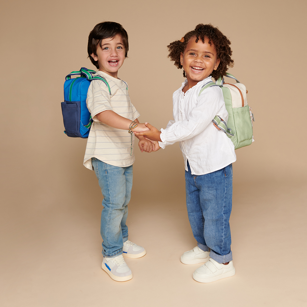 101601544 Itzy Bitzy Bag™ Toddler Backpack - Check Yes! (12) Two young children, each wearing an Itzy Ritzy Sweetie Strap Plus Bear Silicone Pacifier Clip on their backpacks, smile and hold hands while standing against a neutral background.