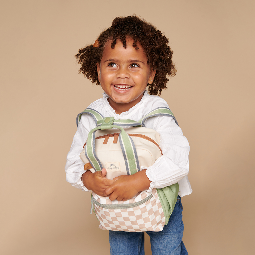 101601544 Itzy Bitzy Bag™ Toddler Backpack - Check Yes! (13) A young child with curly hair smiles while holding an Itzy Ritzy Sweetie Strap Plus Bear Silicone Pacifier Clip. The child is wearing a white long-sleeve shirt and blue jeans, set against a plain beige background.