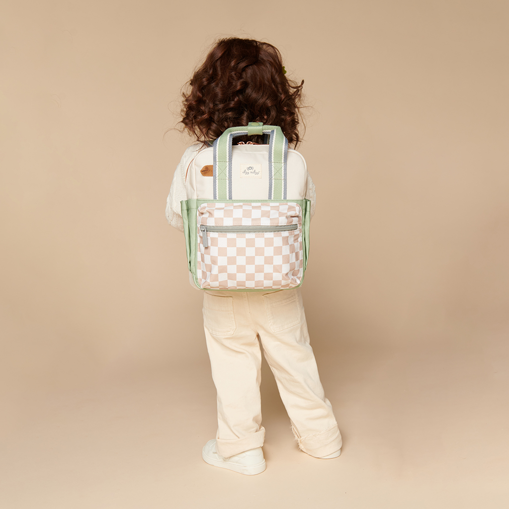 101601544 Itzy Bitzy Bag™ Toddler Backpack - Check Yes! (14) A child with long hair, facing away, stands against a beige background wearing a cream outfit and an Itzy Ritzy Sweetie Strap Plus Bear Silicone Pacifier Clip on their checked backpack with green straps.