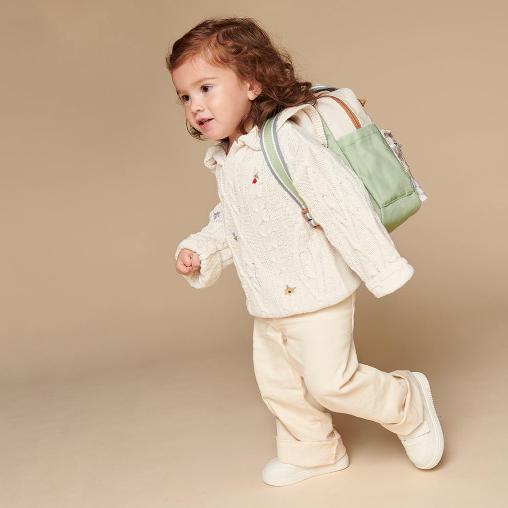 101601544 Itzy Bitzy Bag™ Toddler Backpack - Check Yes! (15) A young child with curly hair wears a beige sweater, light pants, and white shoes, carrying a light green backpack and an Itzy Ritzy Sweetie Strap Plus Bear Silicone Pacifier Clip against a neutral background.