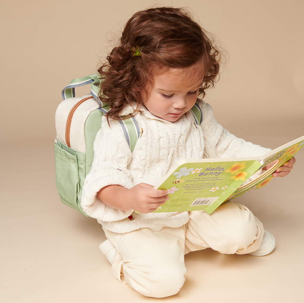 101601544 Itzy Bitzy Bag™ Toddler Backpack - Check Yes! (16) A young child with curly hair, wearing a white sweater and light-colored pants, sits on the floor reading a book titled "Hello Bunny" while sporting an Itzy Ritzy Sweetie Strap Plus Bear Silicone Pacifier Clip.