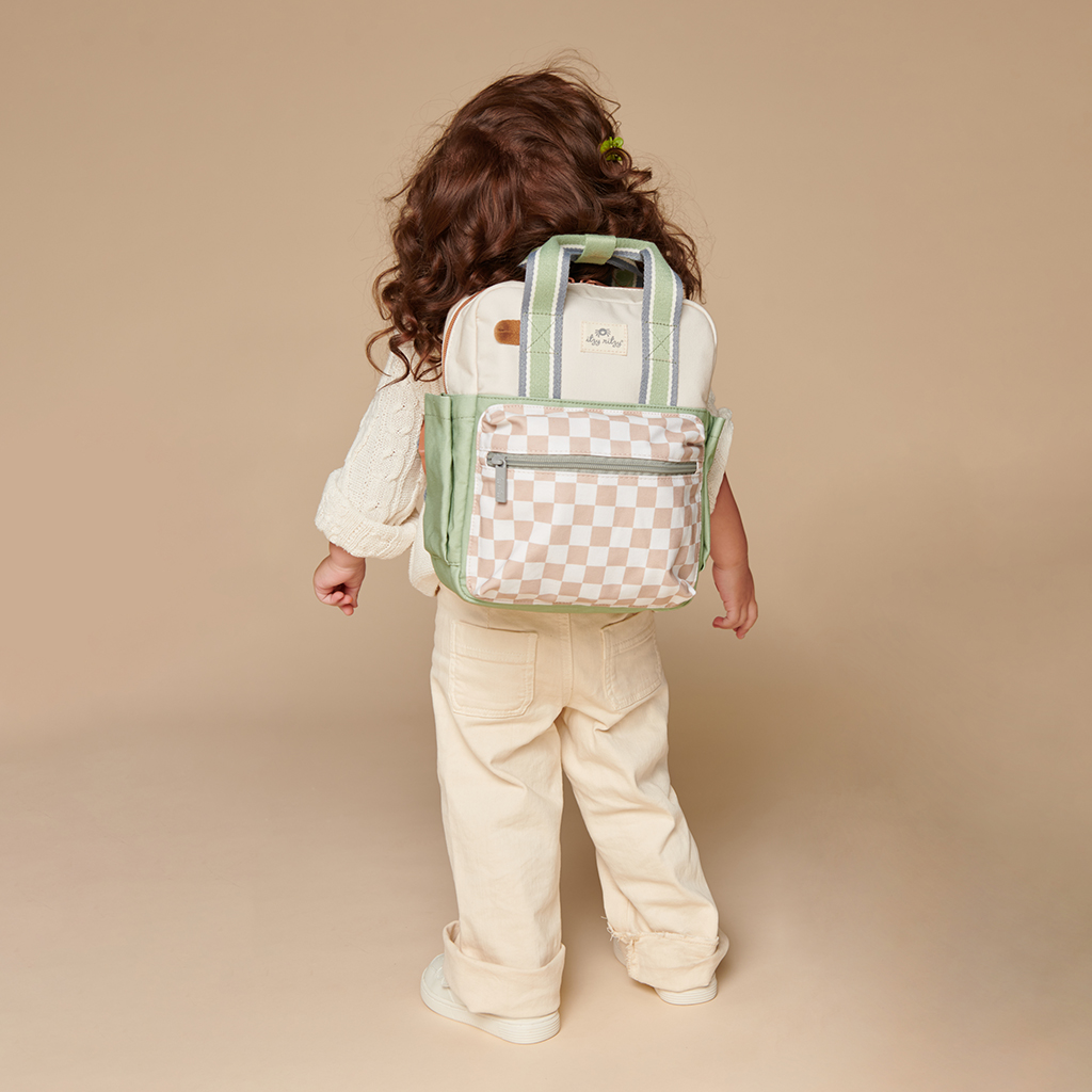 101601544 Itzy Bitzy Bag™ Toddler Backpack - Check Yes! (17) A child with long, curly hair stands facing away, wearing a Itzy Ritzy Sweetie Strap Plus Bear Silicone Pacifier Clip, featuring a large backpack with a checkered front pocket in cream and green colors. The child is dressed in light-colored casual clothing against a plain background.