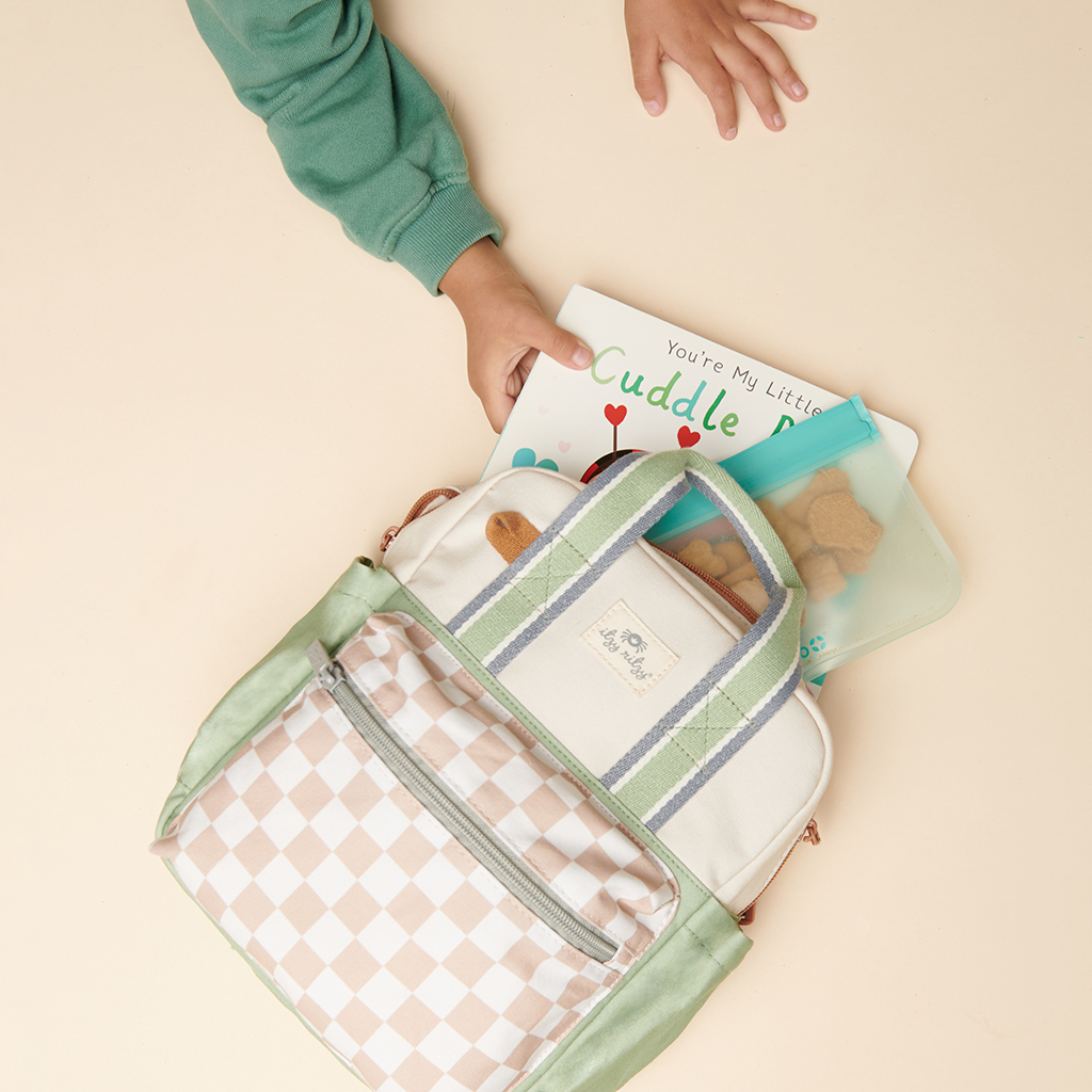 101601544 Itzy Bitzy Bag™ Toddler Backpack - Check Yes! (3) A child reaches for a book from a backpack containing a book titled "Cuddle Bug" and an Itzy Ritzy Sweetie Strap Plus Bear Silicone Pacifier Clip on a light-colored surface.