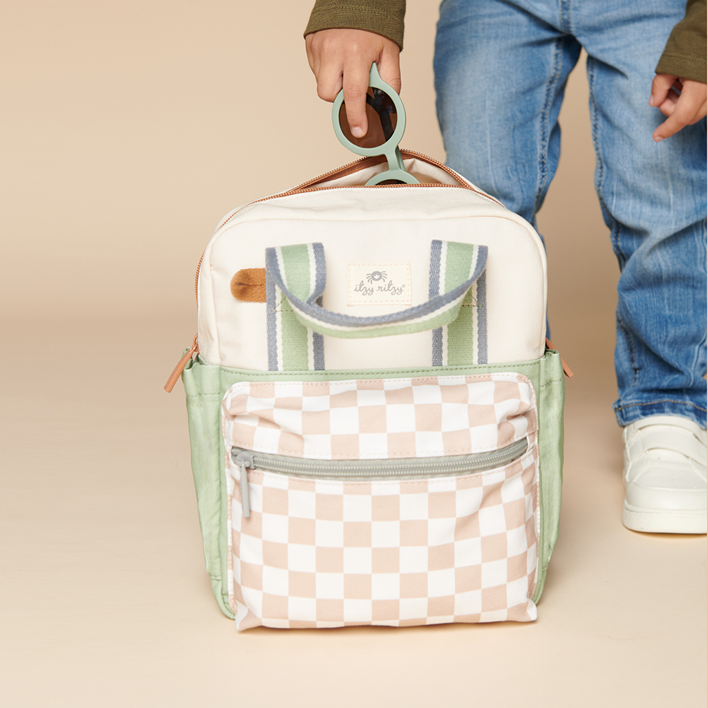 101601544 Itzy Bitzy Bag™ Toddler Backpack - Check Yes! (5) A child holds an Itzy Ritzy Sweetie Strap Plus Bear Silicone Pacifier Clip, featuring a checkered front pocket, green side accents, and striped handles. The background is beige.