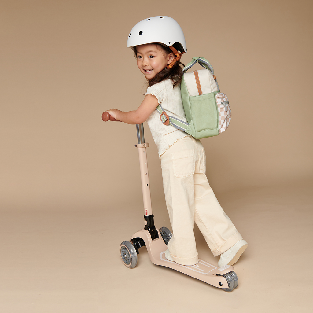 101601544 Itzy Bitzy Bag™ Toddler Backpack - Check Yes! (7) A young child wearing a helmet and backpack rides a three-wheeled scooter on a plain background, with an Itzy Ritzy Sweetie Strap Plus Bear Silicone Pacifier Clip attached to their outfit.