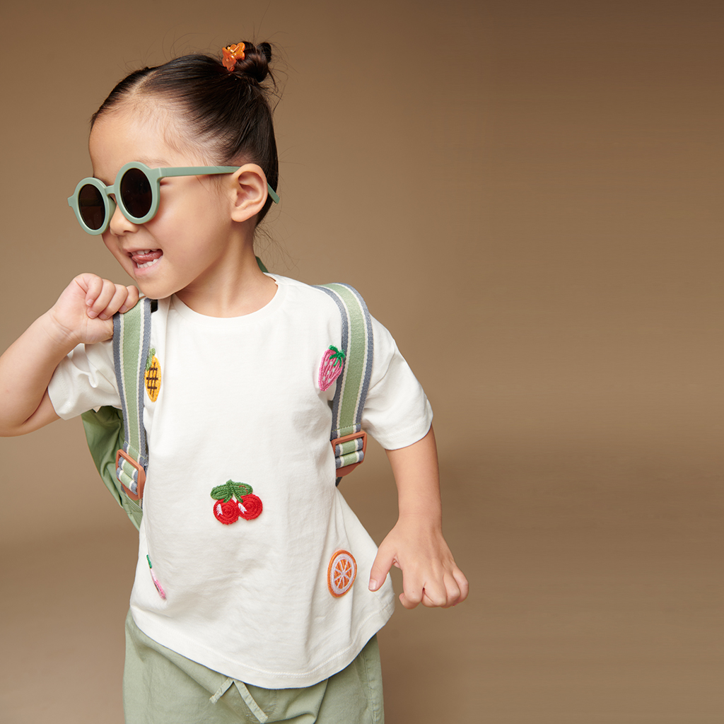 101601544 Itzy Bitzy Bag™ Toddler Backpack - Check Yes! (8) A child with a ponytail and sunglasses smiles while wearing a white T-shirt with fruit designs, green shorts, a backpack, and sporting the Itzy Ritzy Sweetie Strap Plus Bear Silicone Pacifier Clip.