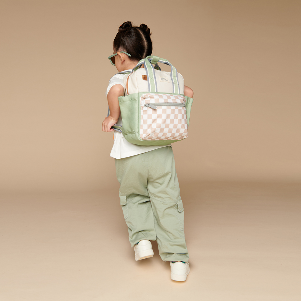101601544 Itzy Bitzy Bag™ Toddler Backpack - Check Yes! (9) A child with two buns in their hair, dressed in a white T-shirt, mint green pants, and white sneakers, is seen carrying the Itzy Ritzy Sweetie Strap Plus Bear Silicone Pacifier Clip on their mint green and checkered tote backpack over their shoulder as they face away from the camera.