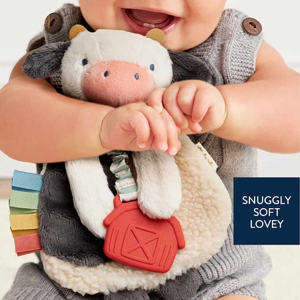 231130_Itzy_Lovey_Cow_Digital_Shelf_Template A baby holds a plush cow toy with colorful tags and a red barn-shaped attachment—text reads "SNUGGLY SOFT LOVEY.