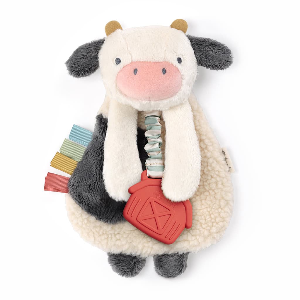 231130_Itzy_Lovey_Cow_Digital_Shelf_Template A plush toy in the shape of a cow with black and white fur, a pink udder, and a red barn-shaped teether attached. Several multicolored fabric tags are also attached to the toy.