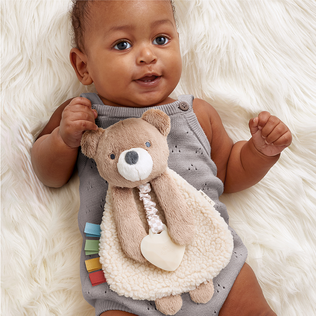 302801667_Itzy_Ritzy_Itzy_Lovey_Bear A baby in a gray outfit lies on a fluffy white surface, holding a plush bear toy with a heart-shaped pacifier.