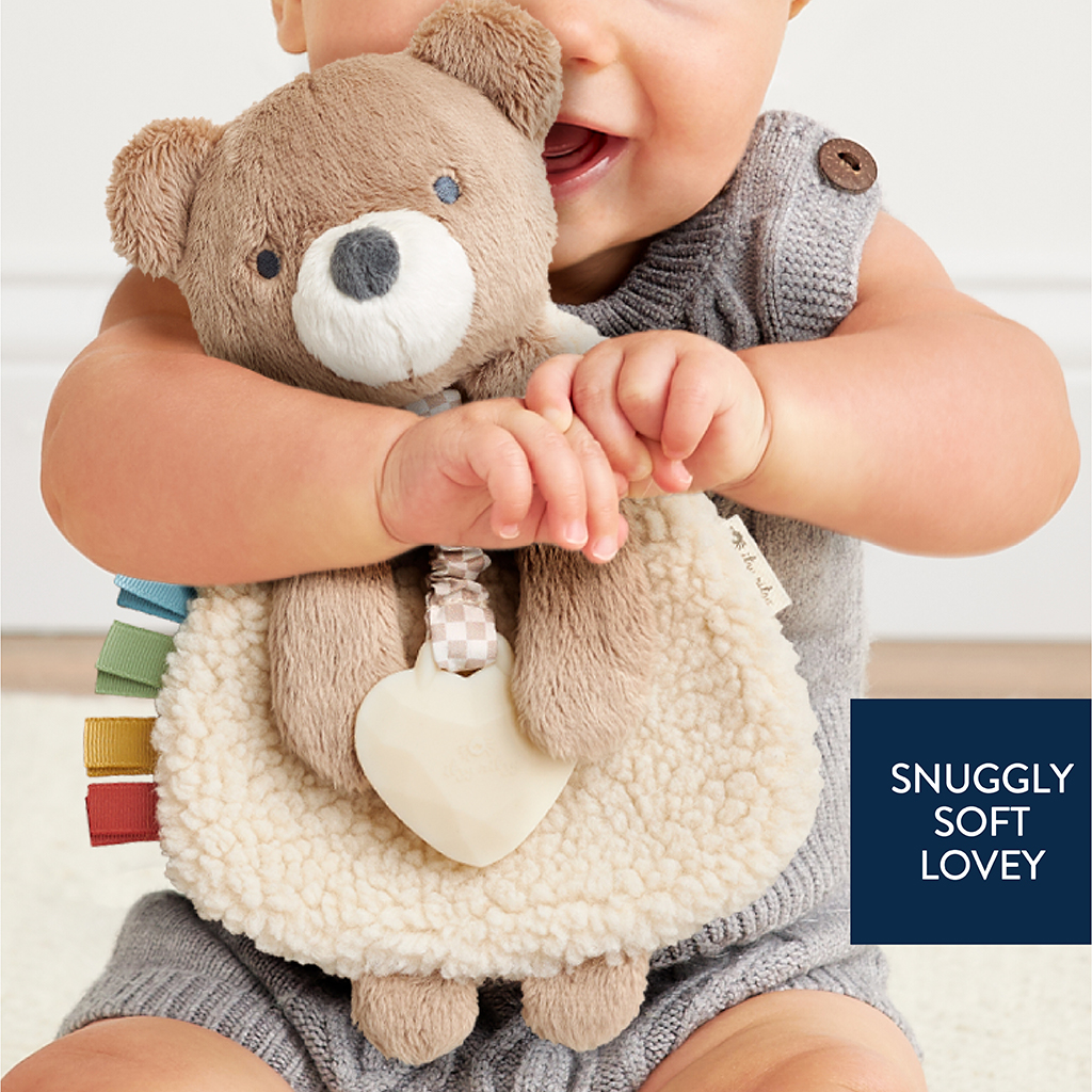 302801667_Itzy_Ritzy_Itzy_Lovey_Bear A baby holds a teddy bear lovey with a soft blanket and attached heart-shaped teether. The text reads, "SNUGGLY SOFT LOVEY.