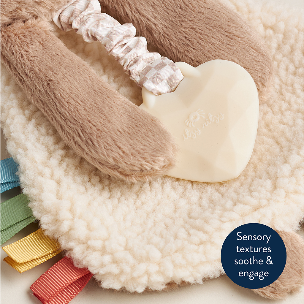 302801667_Itzy_Ritzy_Itzy_Lovey_Bear A soft, textured baby toy featuring a plush fabric, a heart-shaped teether, and colorful ribbon tags, with a text bubble that reads “Sensory textures soothe & engage.”.