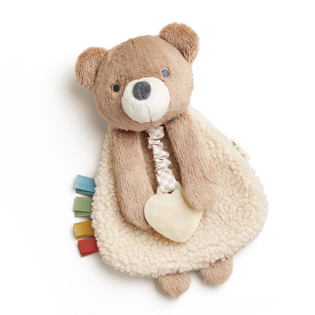 302801667_Itzy_Ritzy_Itzy_Lovey_Bear A soft teddy bear plush toy with a white blanket-like body, holding a heart-shaped teething ring. The toy has colorful ribbon tags attached at the side.