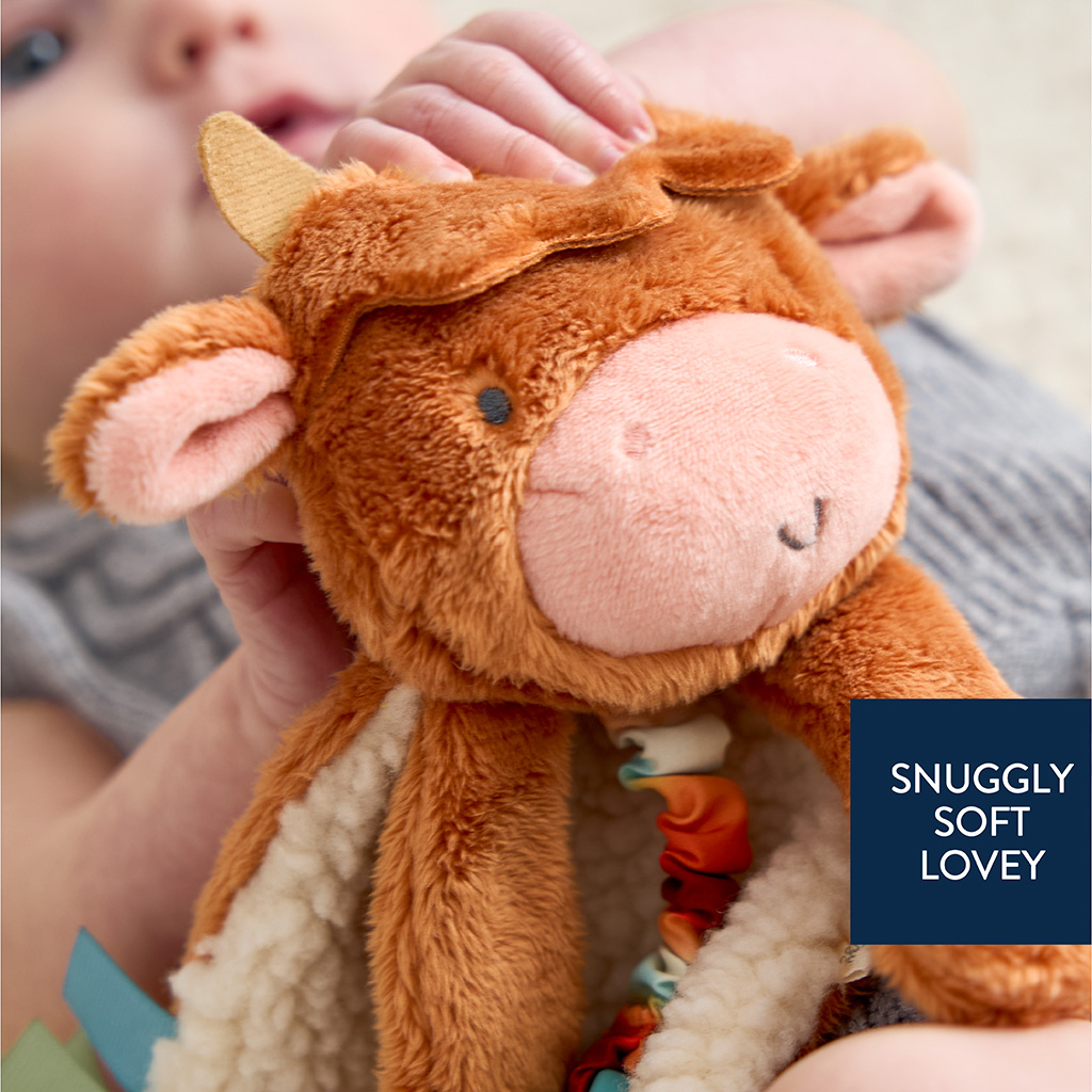 302801668_Itzy_Ritzy_Itzy_Lovey_Highland_Cow A baby holds a soft, plush cow toy labeled "SNUGGLY SOFT LOVEY.
