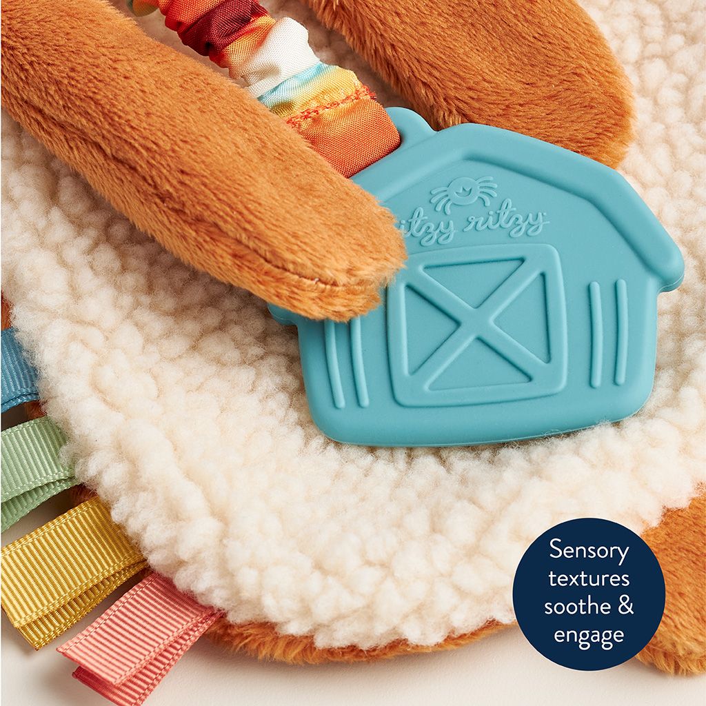 302801668_Itzy_Ritzy_Itzy_Lovey_Highland_Cow Close-up of a sensory toy with various textures, including a soft white fabric, colorful ribbons, and a blue rubber barn-shaped piece with "Itzy Ritzy" text. Round label reads: "Sensory textures soothe & engage.