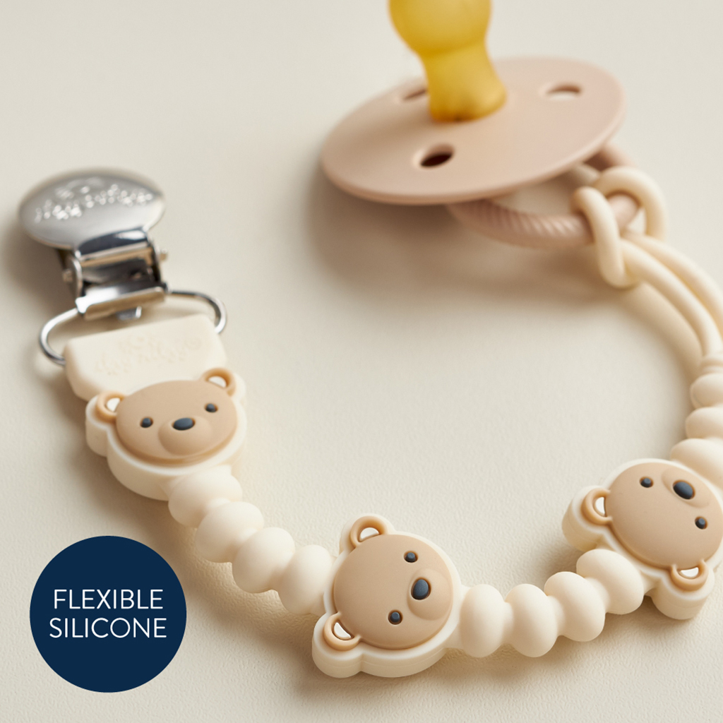 705101559 Sweetie Strap Plus™ Bear (2) The Itzy Ritzy Sweetie Strap Plus Bear Silicone Pacifier Clip in beige, featuring a bear-themed silicone bead clip, is lying on a light surface with a label that reads "Flexible Silicone.