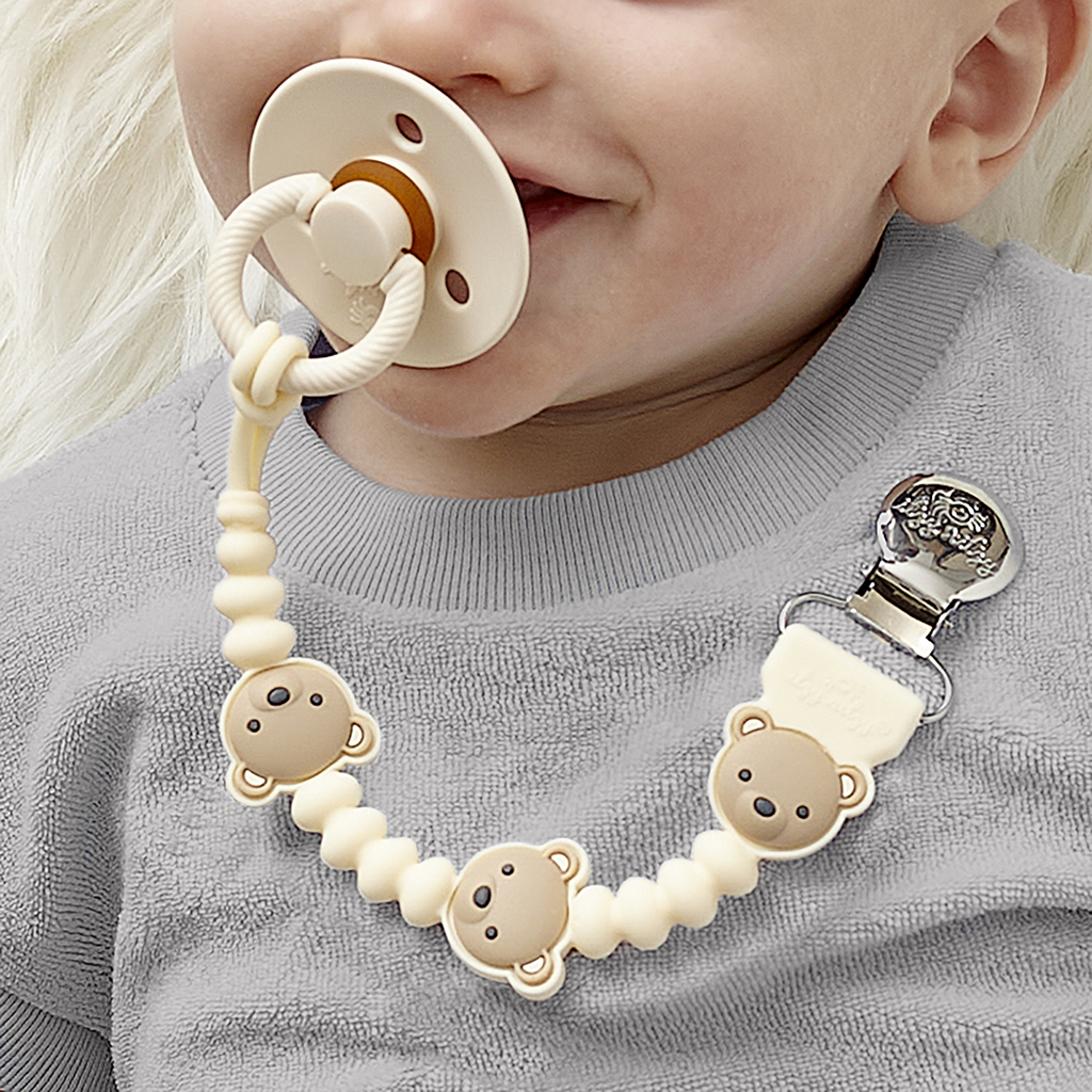 705101559 Sweetie Strap Plus™ Bear (4) A baby with an Itzy Ritzy Sweetie Strap Plus Bear Silicone Pacifier Clip attached to their gray shirt, featuring bear-shaped beads linking to the pacifier.