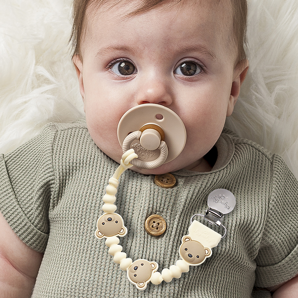 705101559 Sweetie Strap Plus™ Bear (5) A baby with an Itzy Ritzy Sweetie Strap Plus Bear Silicone Pacifier Clip attached to their clothing. The baby is wearing a knitted green outfit with wooden buttons and lying on a fluffy surface.