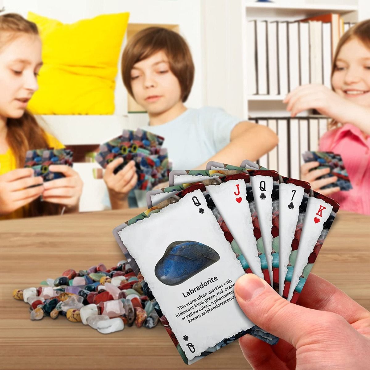716p5sjbosl._ac_sl1500_ Three children playing the National Geographic Rock And Mineral Card Game Gemstone BoardGame Kids Learn STEM Educational NGEO NatGeo. The foreground shows a hand holding a set of cards, one of which features information about labradorite. There is a pile of game pieces on the table.