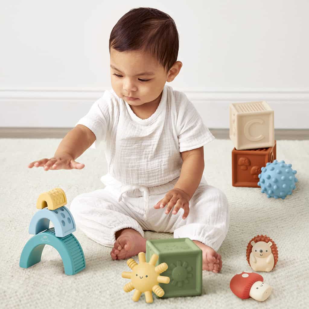 BL853201_Itzy_Ritzy_Toy_Itzy_Blocks_DigitalShelf A toddler in white clothing sits on a carpet, playing with Itzy Ritzy Sensory Blocks 10-Piece Set and a toy hedgehog.