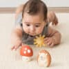 Baby lying on stomach reaching for the Itzy Ritzy Sensory Blocks 10-Piece Set with a plush sun and hedgehog nearby on a carpeted floor.