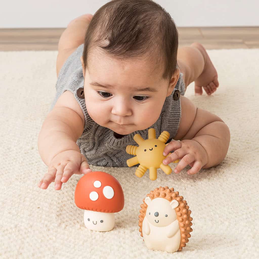 BL853201_Itzy_Ritzy_Toy_Itzy_Blocks_DigitalShelf Baby lying on stomach reaching for the Itzy Ritzy Sensory Blocks 10-Piece Set with a plush sun and hedgehog nearby on a carpeted floor.