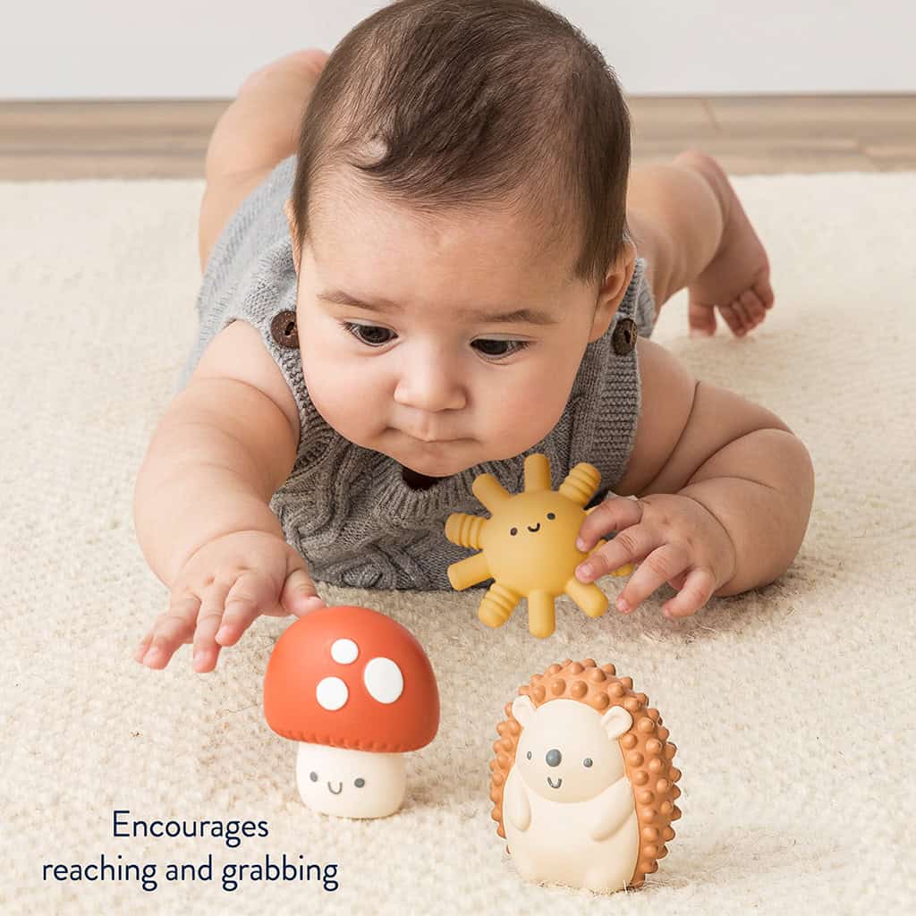 BL853201_Itzy_Ritzy_Toy_Itzy_Blocks_DigitalShelf Infant lying on stomach on a carpet, reaching towards an Itzy Ritzy Sensory Blocks 10-Piece Set and a toy hedgehog.