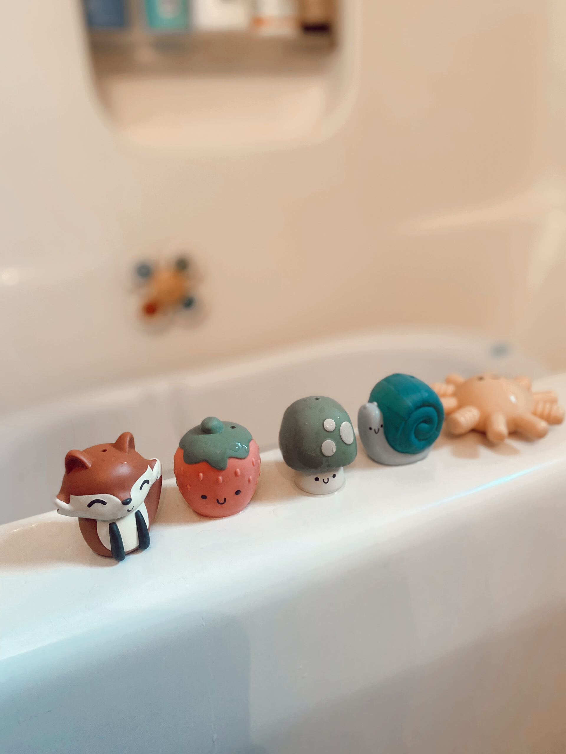 FP8560 Mushroom, Fox, Strawberry, Snail and Sun Squirter (1) Five vibrant Itzy Ritzy Finger Puppets Bath Toys, representing animals and fruits such as a fox, strawberry, mushroom, snail, and sun, lined up on the edge of the bathtub.