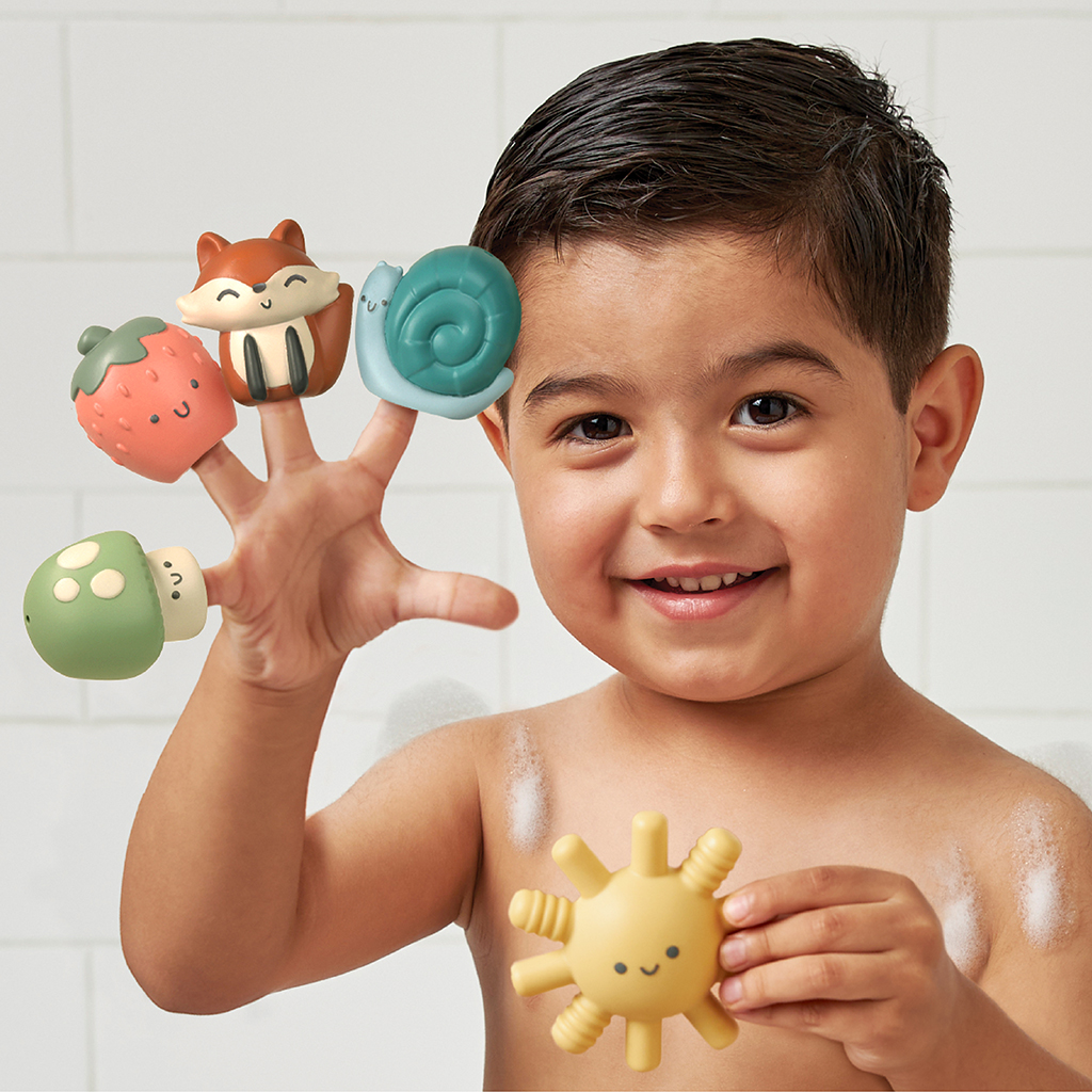 FP8560_Itzy_Ritzy_Bath_Itzy_Bitzy_Finger_Puppets A young child with wet hair stands in a tiled space, holding various Itzy Ritzy Finger Puppets Bath Toys on their fingers and one in the other hand.